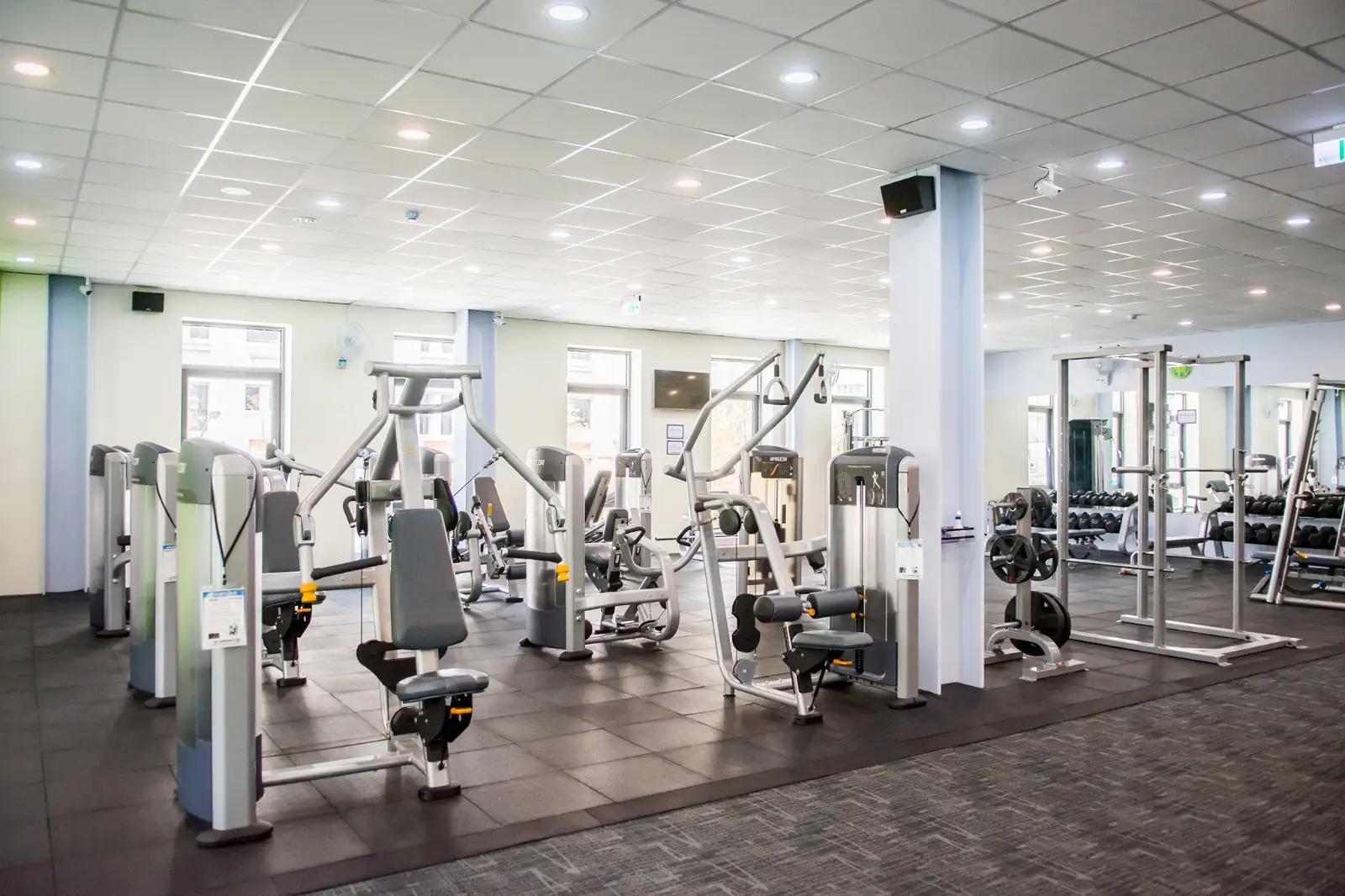 A gym floor with weight machines, squat racks and a dumbbell rack under bright lights